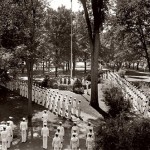 Then: (July) Attending Bates under the World War II V-12 program, Navy men in dress whites stand at attention for morning colors on the Quad. The program, which provided officer training and accelerated college instruction, had some 340 participants at Bates by 1945.  Photograph by George French, 1940s, gelatin silver print, the Edmund S. Muskie Archives and Special Collections Library.