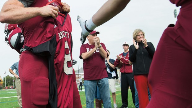 Bates players touch the No. 8 jersey of the late Troy Pappas ’16 while entering Garcelon Field for the Williams game, as Pappas’ uncle Gary Blackwell (Bates shirt) and President Spencer (black coat) show their support. Photograph by Mike Bradley