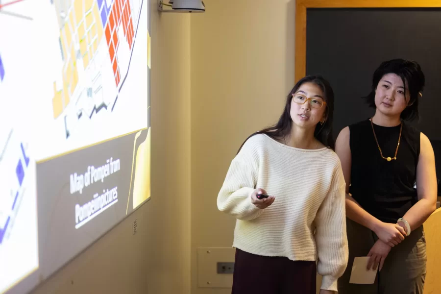 Two students glance at the projector screen while giving a presentation on Pompeii to their class.