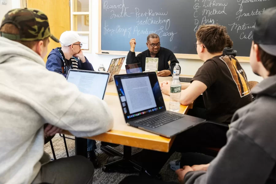 Four students and their professor engage in a small group discussion facing the blackboard in a Pettengill Hall study lounge.