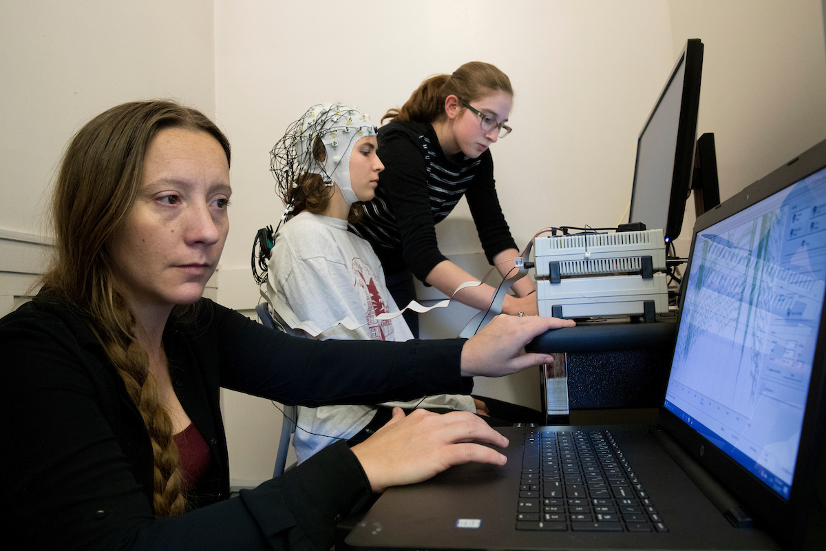 “We are piloting the experiment for these students’ thesis experiments. They were piloting Hannah’s experiment. She’s interested in looking at the extent to which visual masking actually inhibits perception. So when you take a visual mask, you take an image followed by another image, you’re impaired at understanding the first image. The question is why. So what we’re going to do is take the neural activity that we’re measuring. And the nice thing about EEG is that it measures millisecond by millisecond electrical potentials that are generated in the brain , we measure them from the scalp. And we can see over time what the brain is processing and we use machine learning, we put these signals into a computer system tha t reads out the extent to which there is information about what the picture is. We’re wondering, does that information persist when you change the image? Does that persist over time? Hannah’s made the experiment, and we are going to try it out to make sure everything’s ready for participants.”
ν Michelle Greene, assistant professor of neuroscience, says of three thesis students in neuroscience: “They’re all terrific, I might add.”
Hanna De Bruyn ‘18, Old Lyme, Conn. (black striped sweater with glasses)
Katherine “Katie” Hartnett ’18 of St. Paul, Minn. (wearing EEG cap with Bates sweatshirt)
Julie Self ’18 of Redwood City, Calif. (blue plaid shirt)
Email from Hanna: Katie Harnett and I will be testing out our computational neuroscience theses and will be hooking each other up to the EEG tomorrow, Friday, at 12:45-2:30ish in the Bates Computational Vision Lab (Hathorn 108).