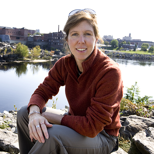 Bates environmental economist Lynne Lewis, associate professor of economics, poses on the Auburn banks of the Androscoggin River behind The Hilton Garden Inn Auburn Riverwatch.Historically, in states like Maine where rivers were treated as part of the industrial infrastructure, pollution and other industrial effects depressed the value of property near rivers. Lewis' Kennebec research offers proof that the opposite effect holds true as well: Restoring a river to a more natural state raises those values.