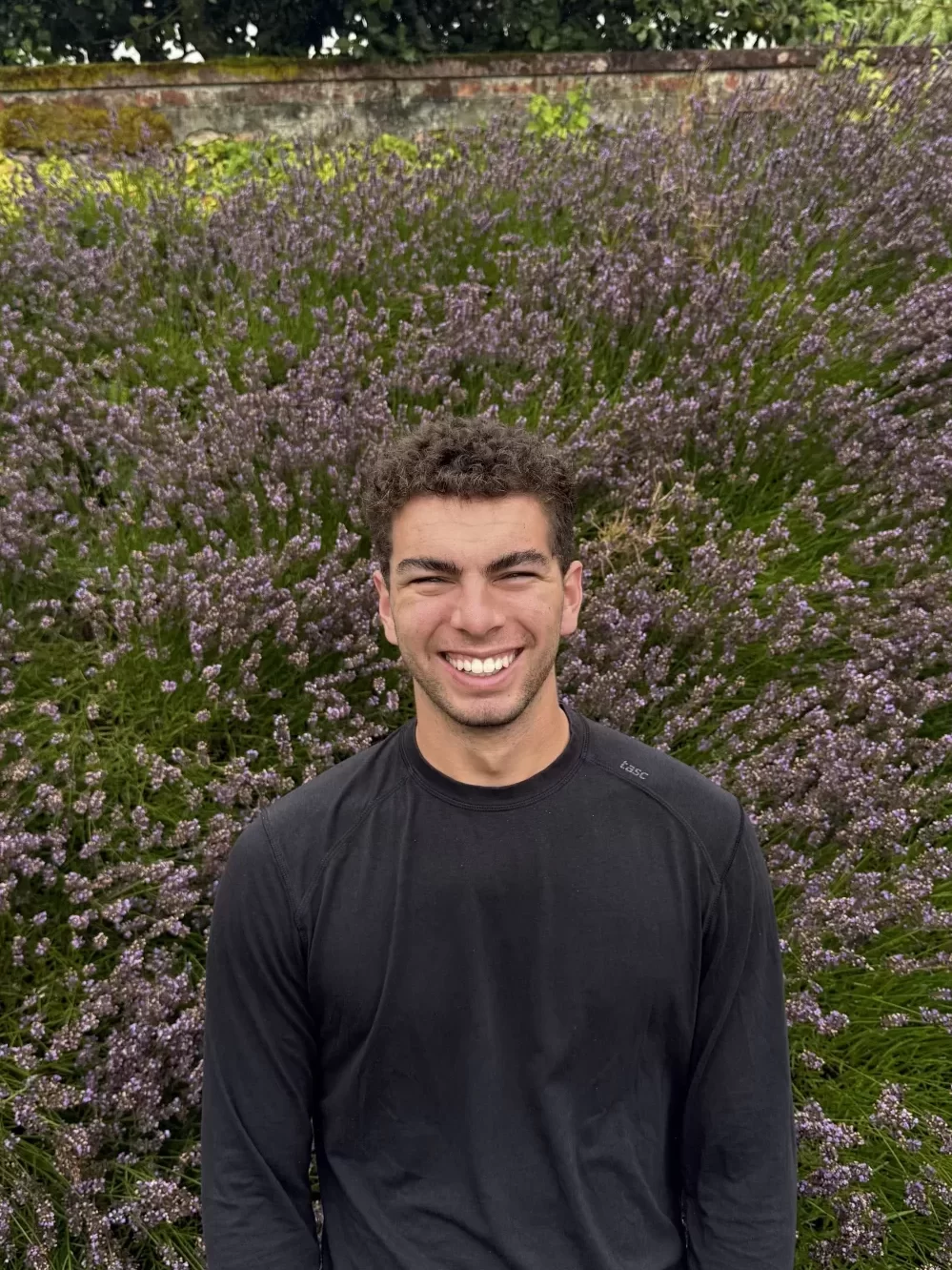 Person smiling outdoors in front of purple flowering bushes, used for a Bates College AESOP profile.