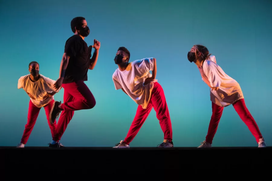 Three dancers watch a fourth dancer move across a backlit stage during a performance.