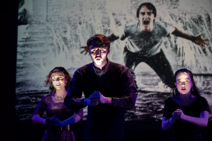 Three students speak on a dark stage, against the backdrop of a large photograph of a man yelling.