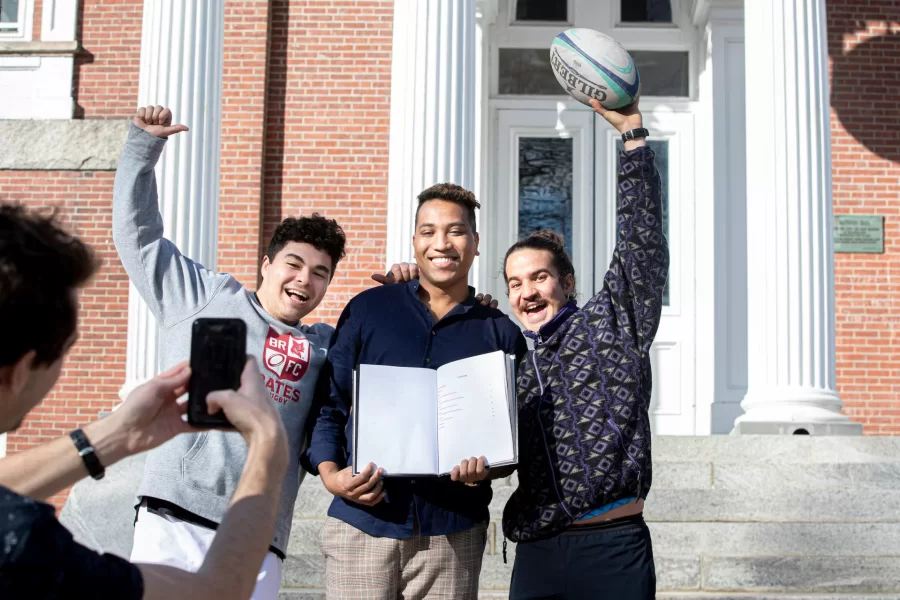 A student poses with his bound senior thesis and two friends in front of Hathorn Hall.