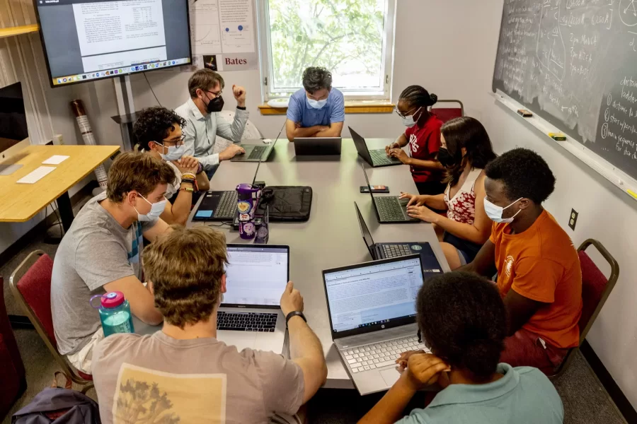 Eight students and one professor seated at a long table in a classroom, looking at data on laptops.
