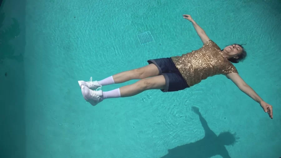 Still image from film "How to Clean a House in 10 Easy Steps) showing an older woman lying in a water with arms outstretched, wearing white sneakers, while crew socks, blue jean shorts, and a gold T-shirt.