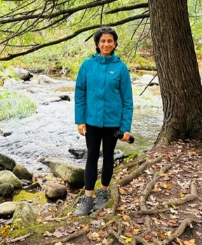 Shreya Arora standing on side of stream, wearing blue jacket, black leggings, and hiking boots