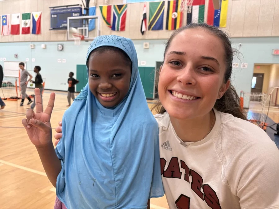 A school-aged child smiling and using hand gesture "Peace" sign standing close and being embraced in a side hug from a smiling college student 
