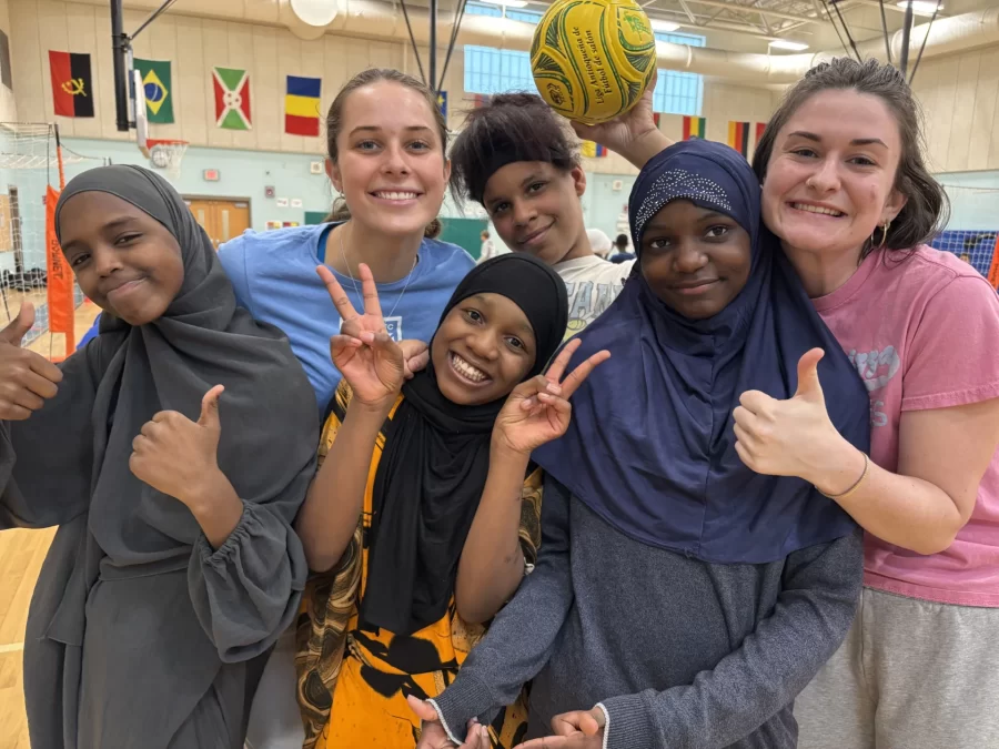 A small group of four school aged children smiling and using hand gestures: "Peace" signs and thumbs up, flanked by two smiling college students