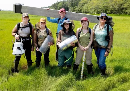A professor stands in a marsh surrounded by five students holding various instruments