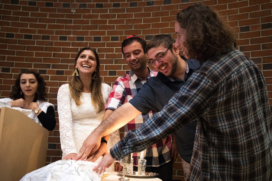 Brooke Drabkin '18 of Roslyn Heights, N.Y., Matthew Winter '18 of New York City, Evan Goldberg '19 of Belmont, Mass., and Alexis Hudes '20 of Owings Mills, Md., place their hands on a loaf of covered challah while listening to Ariel Milan-Polisar, rabbinical student at Hebrew Union College-Jewish Institute of Religion, New York City, left, while in a Shabbat Evening Service & Dinner in the Benjamin Mays Center during Back to Bates Weekend. The Back to Bates Shabbat dinner and service run by Bates Hillel. (Theophil Syslo/Bates College)
