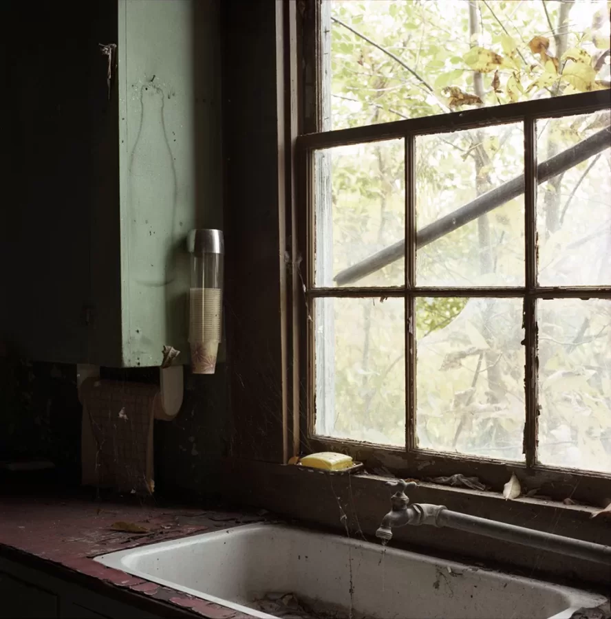 Dimly lit interior scene of a kitchen sink in front of a dingy window, paint peeling on the counter and window wood