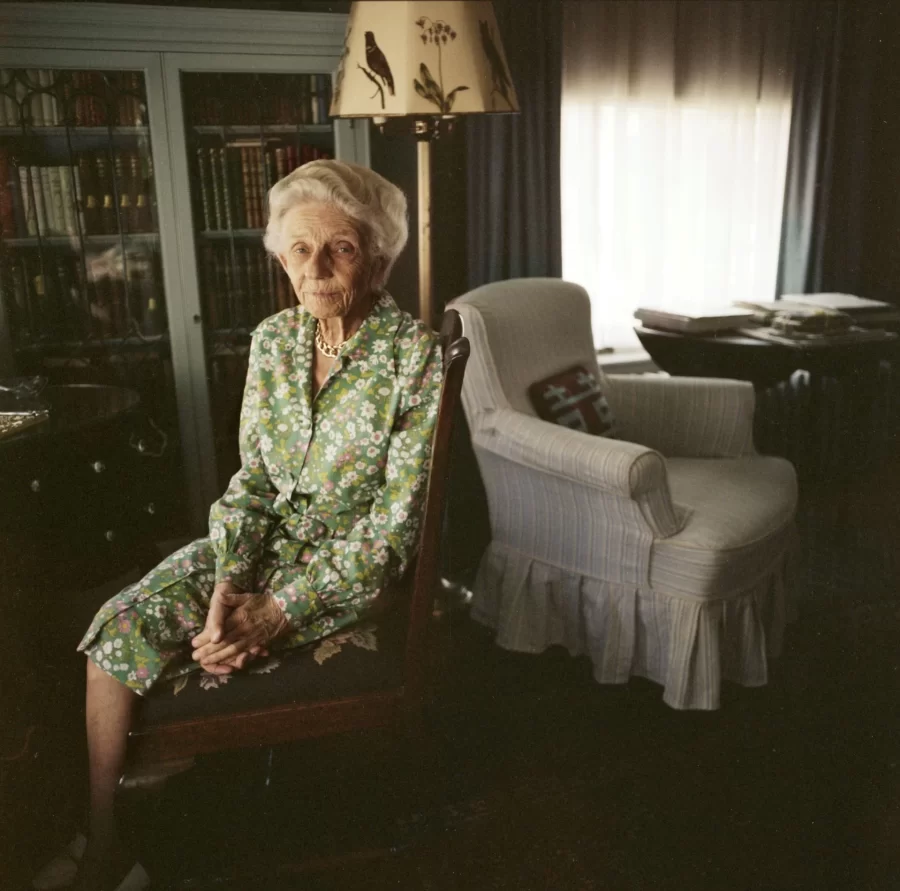An elderly woman with white hair, dressed in a green floral dress, sits in a needlepoint-cushioned wooden chair in a living room
