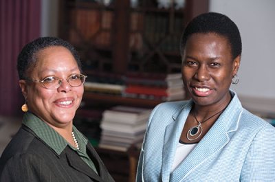 Carmita McCoy (green suit with glasses) and Marylyn Scott (checked blue suit) pose in Lindholm House. They share a position, the Director of Multicultural Recruitment and Associate Dean for Student Transition, known as "swing dean."