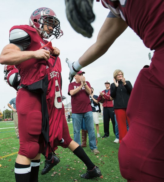 Bates players touch the No. 8 jersey of the late Troy Pappas ’16 while entering Garcelon Field for the Williams game, as Pappas’ uncle Gary Blackwell (Bates shirt) and President Spencer (black coat) show their support. Photograph by Mike Bradley