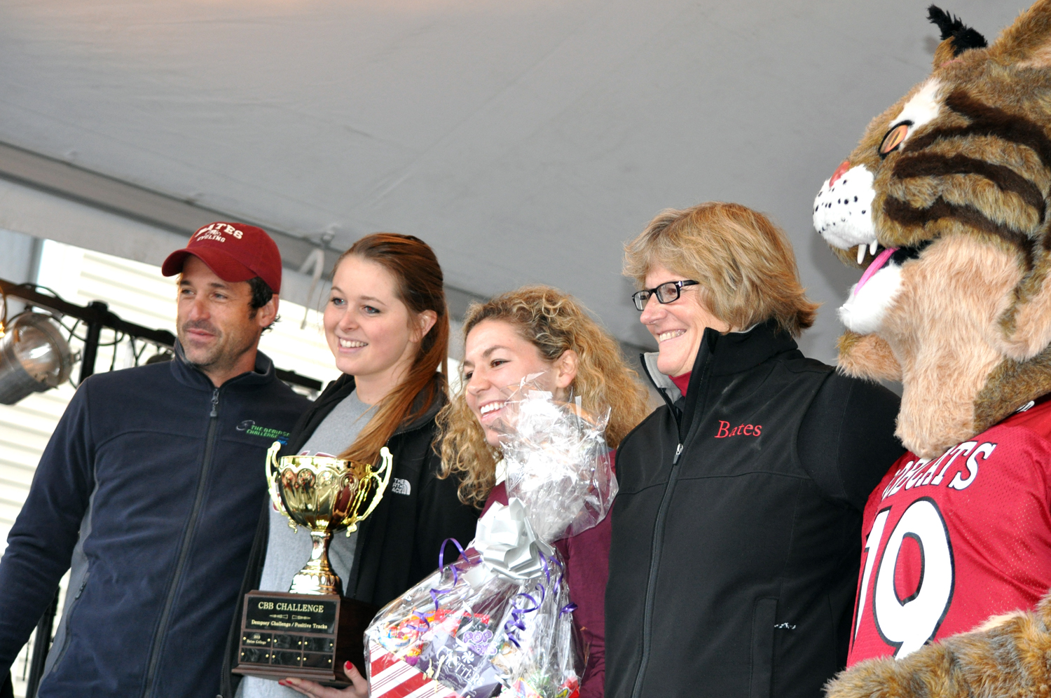 Patrick Dempsey presents the CBB Challenge College Cup to, from left, organizer Hillary Throckmorton '15, Abby Zwetchkenbaum '15 (one of two top Bates fundraisers, along with Julia Gordon '15), President Clayton Spencer and the Bates Bobcat on Oct. 13 during a ceremony in Lewiston's Simard-Payne Park. (Meg Kimmel/Bates College)