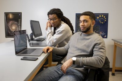 Physics students Kingdell S. Valdez ‘19 of Andover, Mass. (gray sweater) and Cristopher Thompson '19 of Macon, Ga., (Bates track sweatshirt) pose for photographs and discuss a physics problem at the blackboard in third-floor Carnegie Science physics lab on March 7, 2019.They are working on a collaborative physics senior project, doing different aspects w/similar data, turning in separate documents.