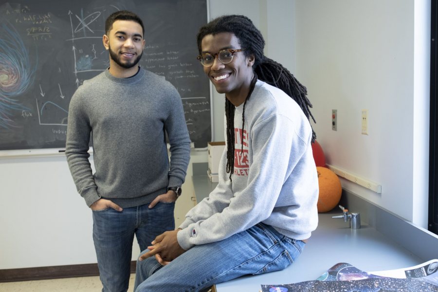 Physics students Kingdell S. Valdez ‘19 of Andover, Mass. (gray sweater) and Cristopher Thompson '19 of Macon, Ga., (Bates track sweatshirt) pose for photographs and discuss a physics problem at the blackboard in third-floor Carnegie Science physics lab on March 7, 2019.They are working on a collaborative physics senior project, doing different aspects w/similar data, turning in separate documents.