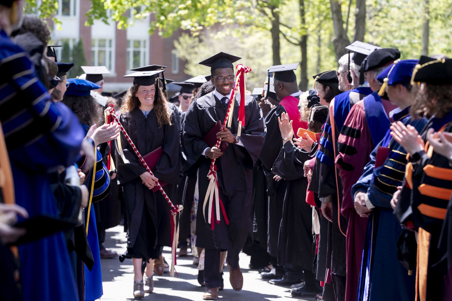 Bates College 2019 Commencement (the one hundred and fifty-third) on the Historic Quad, at which Travis Mills receives an Doctor of Humane Letter. Placing the collar on Mills is the college's mace bearer, Charles Franklin Phillips Professor of EconomicsMichael Murray.Senior class marshals Katherine Gallard and Michael Driscal Jr. lead the student recessional as they pass through two lines of faculty.