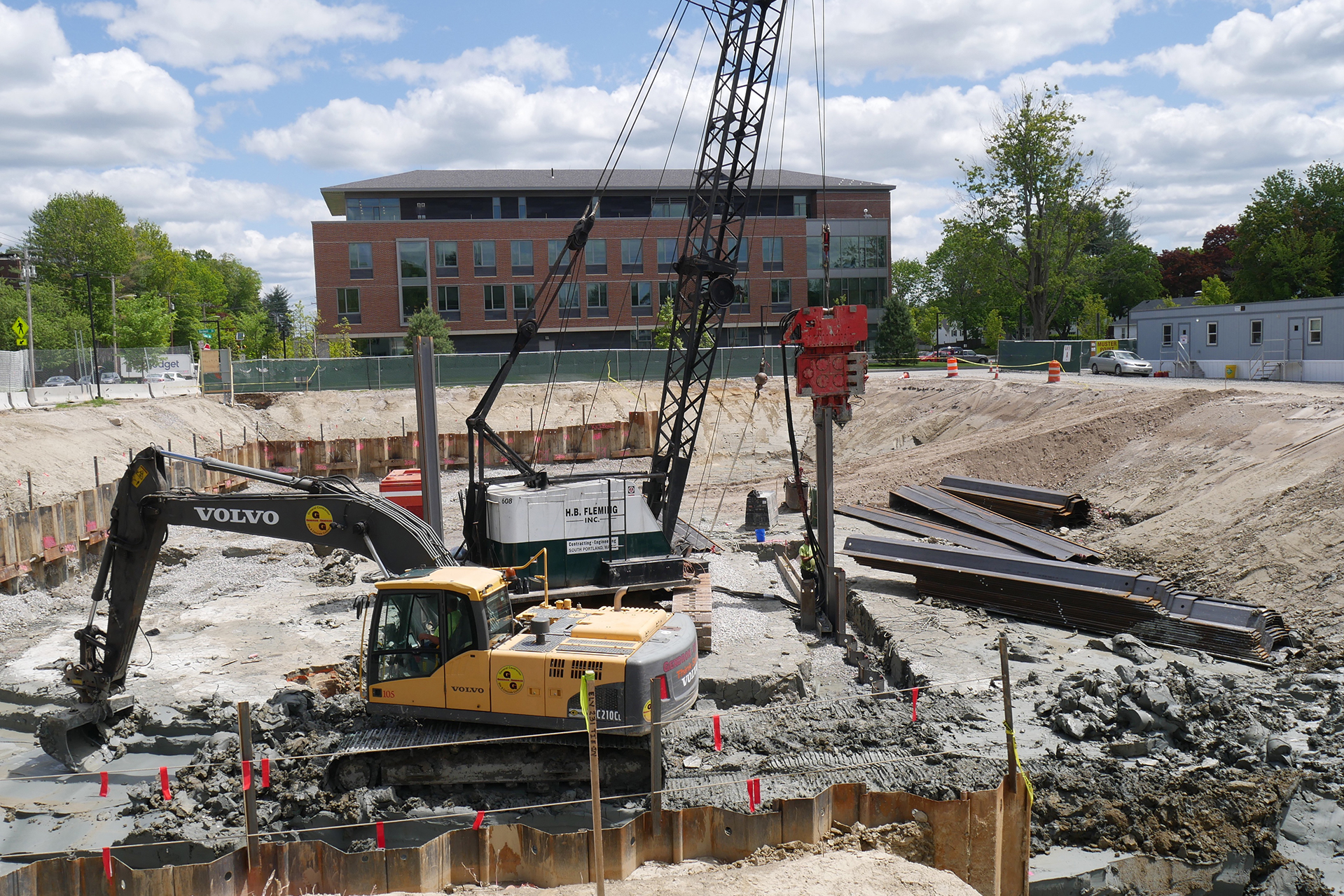 It's full-tilt action in the science building foundation hole on June 4. Suspended from the Fleming Inc. crane, the red vibro hammer is driving a sheet pile at a secondary excavation. Meanwhile, the yellow power shovel is removing clay that it will dump in a heap, not to be confused with a pile, for a second machine to load into a truck. (Doug Hubley/Bates College)