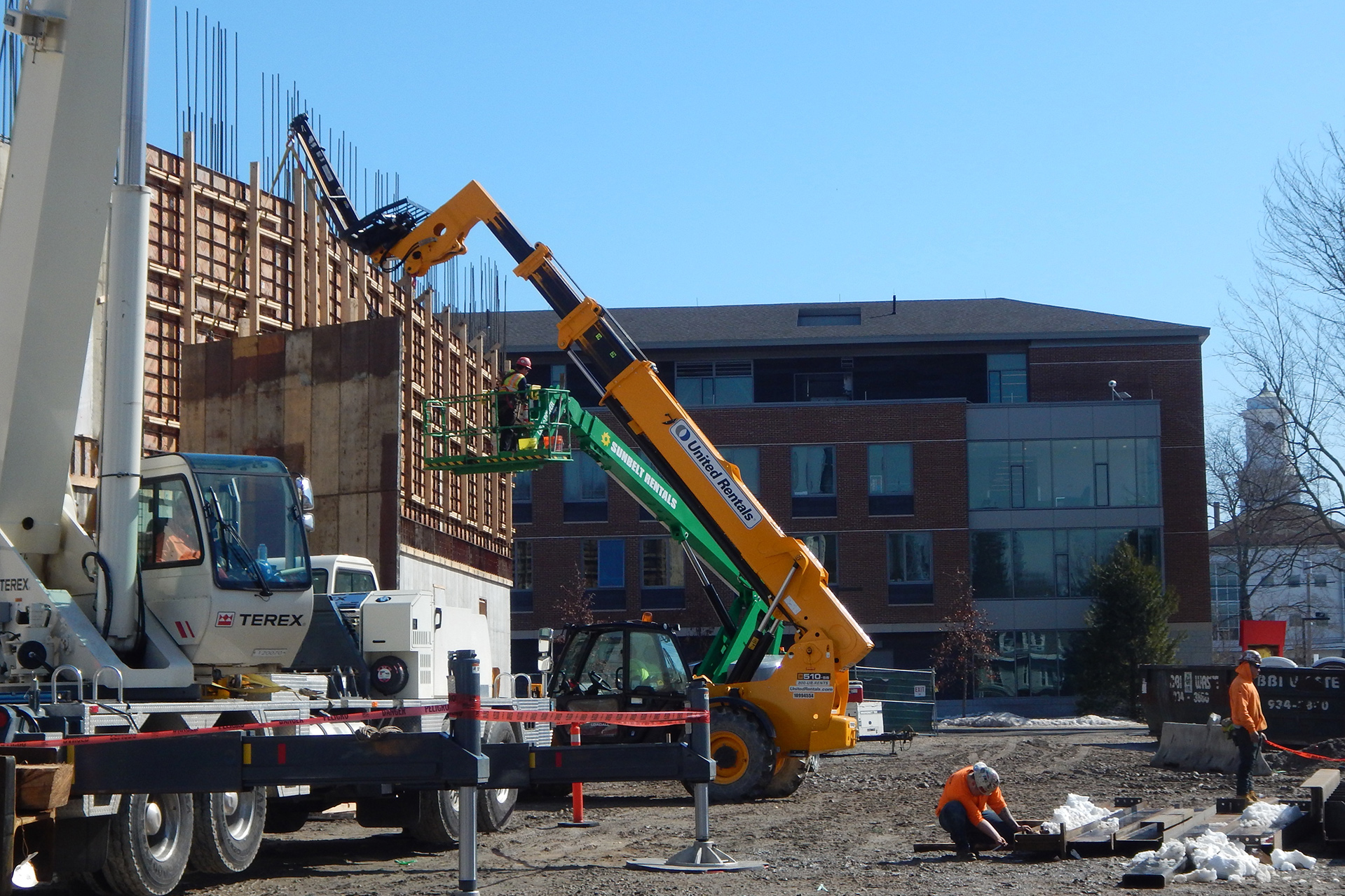 The yellow machine, a Lull-style or telescoping-arm forklift, lowers a concrete form panel from the Bonney Center south wall. After new concrete has cured, the forms can be stripped off and re-used. (Doug Hubley/Bates College)