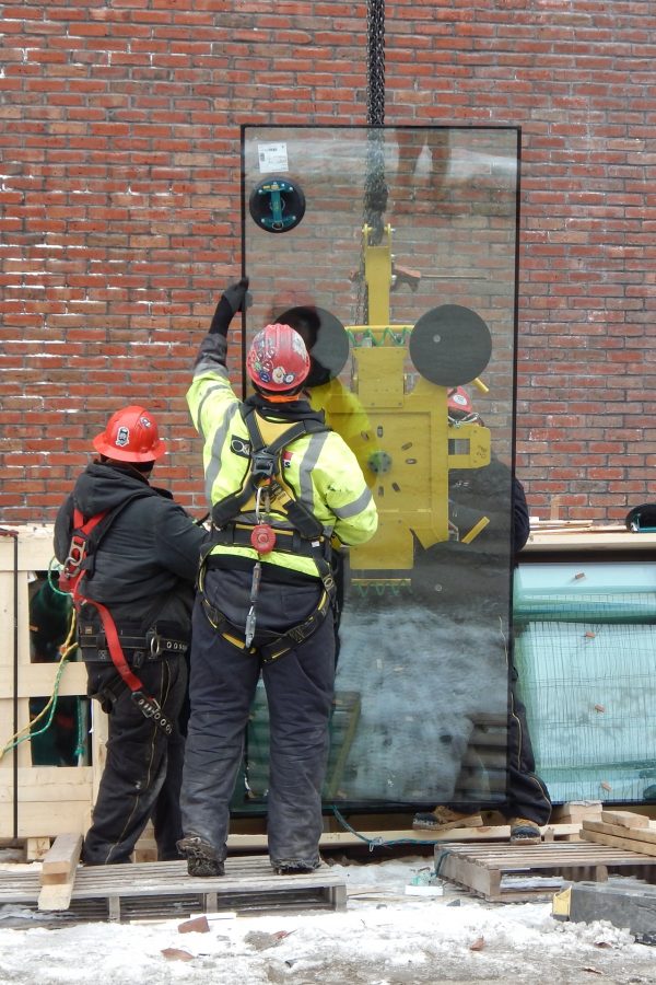 On Feb. 1, a crew from O&P Glass has uncrated a piece of glass for the Monumental Stair curtain wall and fastened the yellow vacuum lifter to it. Next, a glassworker will guide the pane as a lift carries it around the corner to its destination.  (Doug Hubley/Bates College)