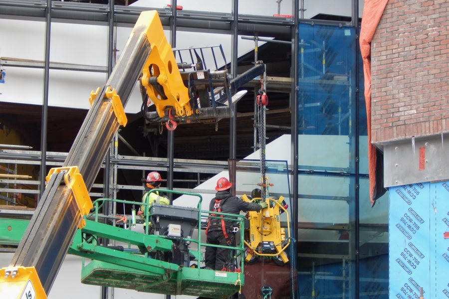 With the help of a vacuum lifter that keeps a tight grip on the heavy pane, workers from O&P Glass maneuver a section of glass into place on the Monumental Stair curtain wall on Feb. 1. To the right of the manlift, pieces installed earlier are reflecting nearby blueskin weather barrier.(Doug Hubley/Bates College)