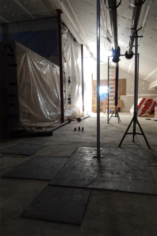 The Hedge Hall–facing end of Dana’s attic. The plastic-wrapped object at left is the HVAC machine installed in late November. At top are steam pipes that will connect to the machine. The steel plates in the foreground cover holes in the concrete floor — because the attic is not a public space, the plates will be given a beveled border for safety reasons but otherwise left as they are. (Doug Hubley/Bates College)