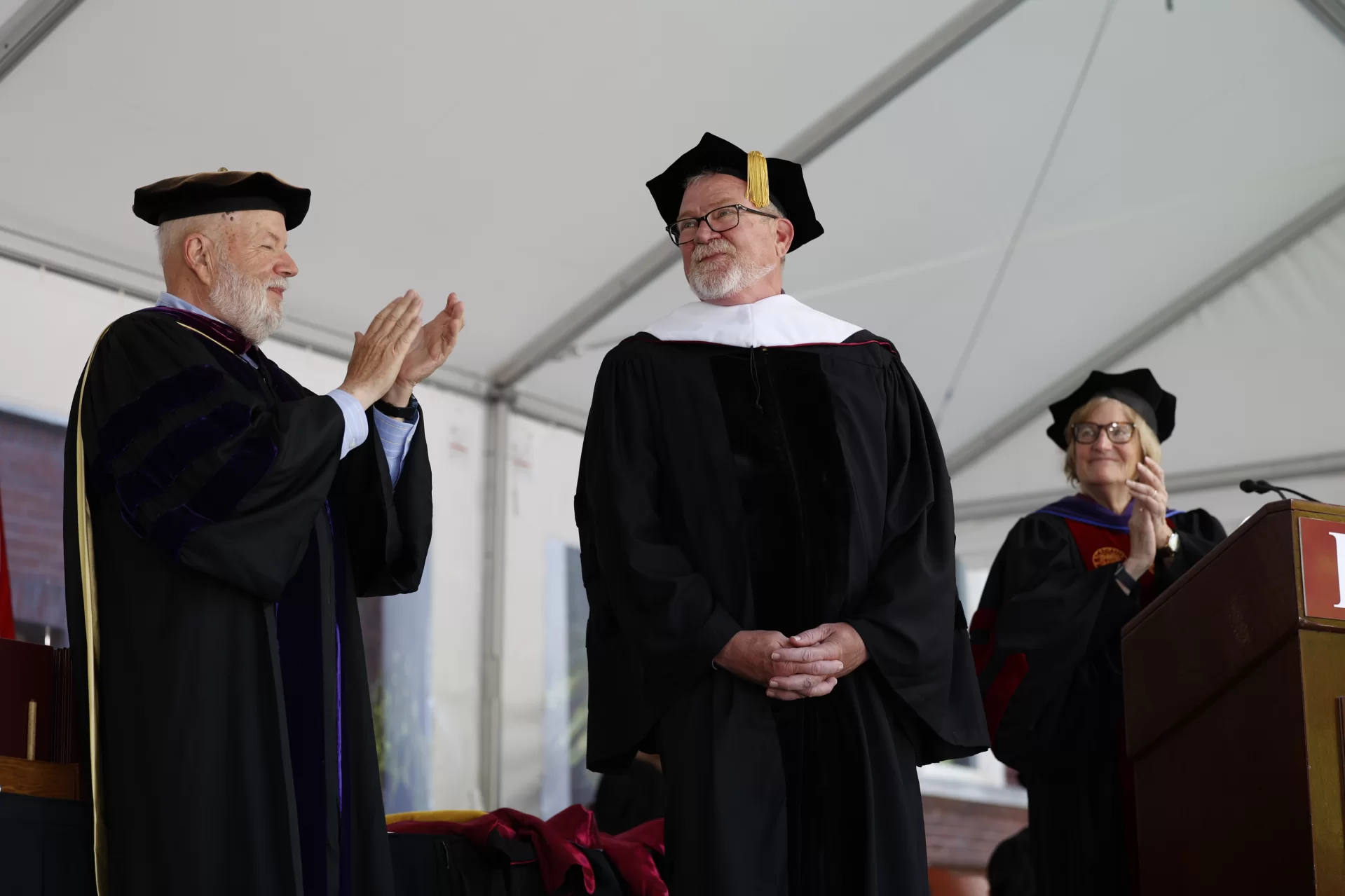 Honorary degree recipient Michael Bonney '80 accepts applause from Michael Murray, Phillips Professor of Economics, and President Clayton Spencer at Bates Commencement on May 29, 2022. (Phyllis Graber Jensen/Bates College)