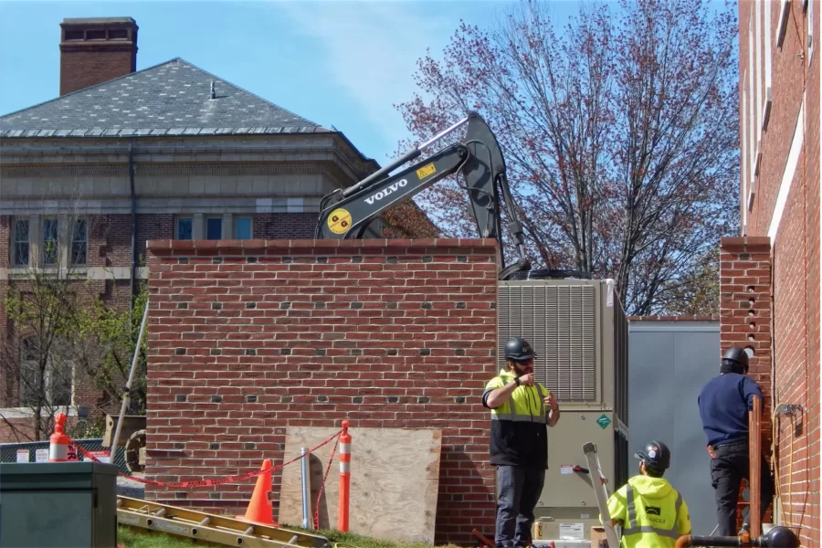 Pipefitters connect Dana Hall’s new chiller, the gray item to the right of center, to the building's interior HVAC network. The man at right is drilling holes in brick. The excavator behind the chiller well is contouring soil for landscaping. Coram Library is in the background. (Doug Hubley/Bates College)