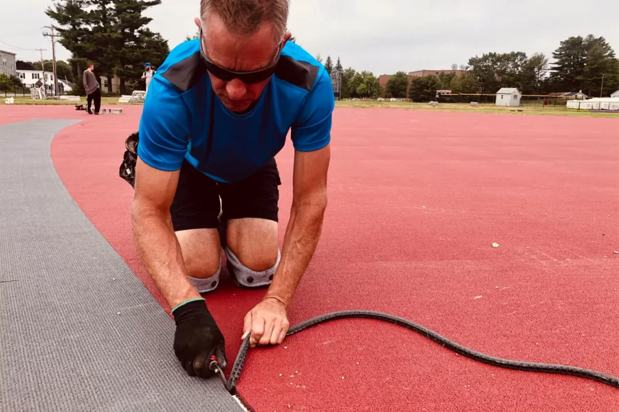 Corey Ernst of Wicked Flooring, the Illinois-based subcontractor laying the Mondo surface on the Russell Street Track, hand-trims an edge to ensure a perfect fit. (Jay Burns/Bates College)