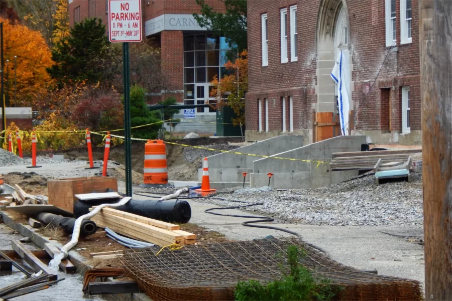 The construction zone in front of Chase Hall contains supplies — lumber, weatherproofing fabric, various forms of rebar, etc. At lower left, a hose drains an excavation near the building. To the right of center are new retaining walls near a building entrance. (Doug Hubley/Bates College)