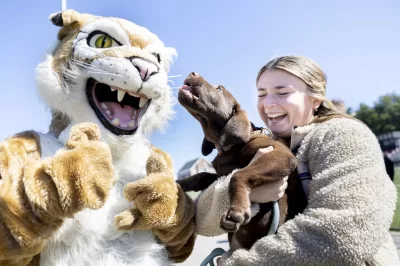 Cats Carnival, held for the first time in two years, was held on the Bardwell Street Field, on Saturday, Sept. 24, 2022.After Two-Year Hiatus, 'Cats Carnival Returns This SaturdayThe fall-themed 'Cats Carnival, hosted by Bates College Athletics, returns after a two-year hiatus this Saturday just before the Bates Football home opener vs. Tufts at 1:30 p.m. on Garcelon Field. The carnival features bounce houses, face painting, poster making, a prize wheel, concessions, and games for kids of all ages. Bates student-athletes will volunteer at the family-friendly event. The 'Cats Carnival is free and open to the public. Bates Athletics invites community members to join the fun and to cheer on the Bobcats in all their home contests. Admission to all games is free.Kirsten Pelletier ’20 and her four-month-old-chocolate Lab Goose enjoy a photo opportunity with the Bates Bobcat.