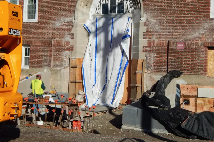 A view of the Campus Avenue entrance to Chase Hall near Kenison Gate. The black blanket in the right foreground is draped over a new retaining wall and precast concrete seating. (The blanket keeps fresh concrete at the right temperature for curing.) The orange "flowers" in the left foreground are safety caps on a patch of rebar for another section of wall. (Doug Hubley/Bates College)