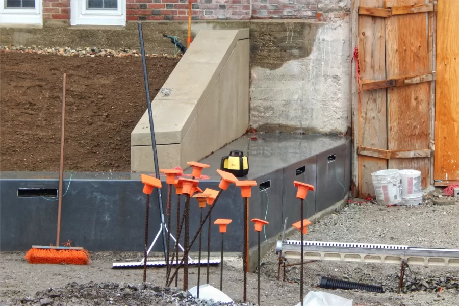 In this view near the main Campus Avenue entrance to Chase Hall, the dark gray masonry is bench seating. The wires will connect to lamps in the openings. The orange-capped rebar is part of footings for steps leading from the sidewalk to the building. At lower right is a drain. (Doug Hubley/Bates College)