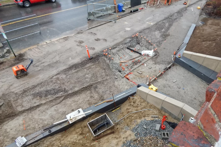 Chase Hall’s main Campus Avenue entrance is shown from a second-floor window. Two accessible ramps converging from left and right will meet a short flight of steps at center, marked by orange rebar caps. The dark gray masonry is bench seating. Low in the image, the gray box just left of center is a drainage vault. (Doug Hubley/Bates College)