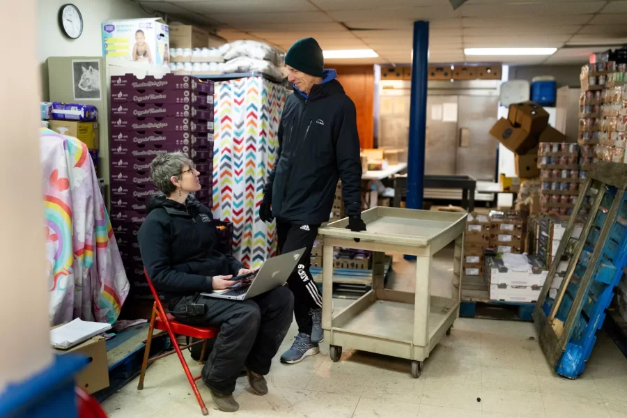 Erin Reed ’08, executive director of the Trinity Jubilee Center in Lewiston, works in the organization’s food bank on Nov. 30, 2023, after which she relocates into the center’s shelter on her computer. Shown with her mentor and retired ED Kim Wetlauffer ’80.