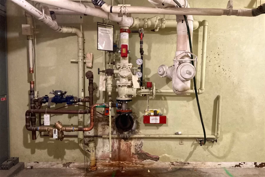 A scene from the basement of 96 Campus Ave.: Through the copper and white-painted apparatus at center, municipal water enters the building and is divided between domestic use and the sprinkler system. To conform with updated codes, the domestic and sprinkler feeds will be renewed and completely serparated as part of the current renovation. (Doug Hubley/Bates College)