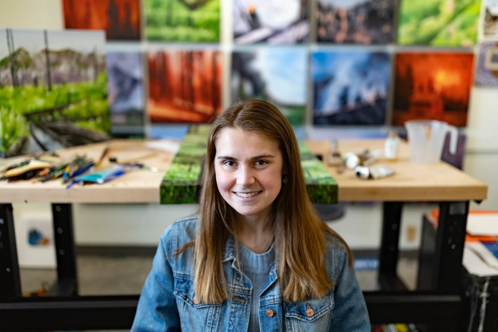 Studio art major Amelia Hawkins ’24 of Sun Valley, Idaho, in her Olin Arts Center studio with her paintings for her senior exhibition.

Artist Statement
“Living in Idaho for most of my life, there has been a common thread which never changes through the years. I consider Idaho to have five seasons: Fall, Winter, Spring, Summer and Smoke. Once August rolls around, the smoke rolls in with it. Forest fires are inevitable here. They have been for centuries, and can actually be good for the cycles of the ecosystem. Fires reduce dead vegetation and stimulate new growth, resulting in overall improvements of wildlife habitats. These good consequences cannot happen if fires occur too frequently. Over my lifetime, the frequency of local fires and smoke coming from fires in California and Canada has increased. Summer camp was often canceled due to unhealthy air quality. I remember asking my mom, “Where are all these ashes coming from?” This has always stuck with me, and I wanted to show this aspect of my life in my current work.

Working in oil on square and rectangular canvases, I paint scenes representing the different stages of forest fires. The scenes with bright flames reflect the early stage when the fire has just started. Days, weeks, or months later, the fire will go out. It will reveal scenes of barren forest, with hazy ash and smoke. Winter will soon come when glistening snow falls all around the dead trees, creating patterns of blue shadows on mountain faces. The snow will start melting in May and regrowth will begin. A lush landscape, full of lupin flowers, will emerge at the base of the dark burned trees. It is an amazing cycle which captivates me every year.

I am interested in the juxtaposition of the destructive chaos of the early stages and serenity of the
later stages. Given my love for pattern design, I knew I wanted to create a pattern conveying these different environments. With that intention, I painted a variety of colorful scenes in identical square and rectangular formats that would then be unified within the arrangement of a grid. To further my vision, I was also inspired by the work of Philip Juras who depicts forest fires in his paintings. He often works from direct observation, painting fire adapted landscapes of the Southeast. Like Juras, I experiment painting with faster brushstrokes to evoke the essence of a fire in real time. I hope my work provides a glimpse of forest fires and the consequences they bring to our world.”