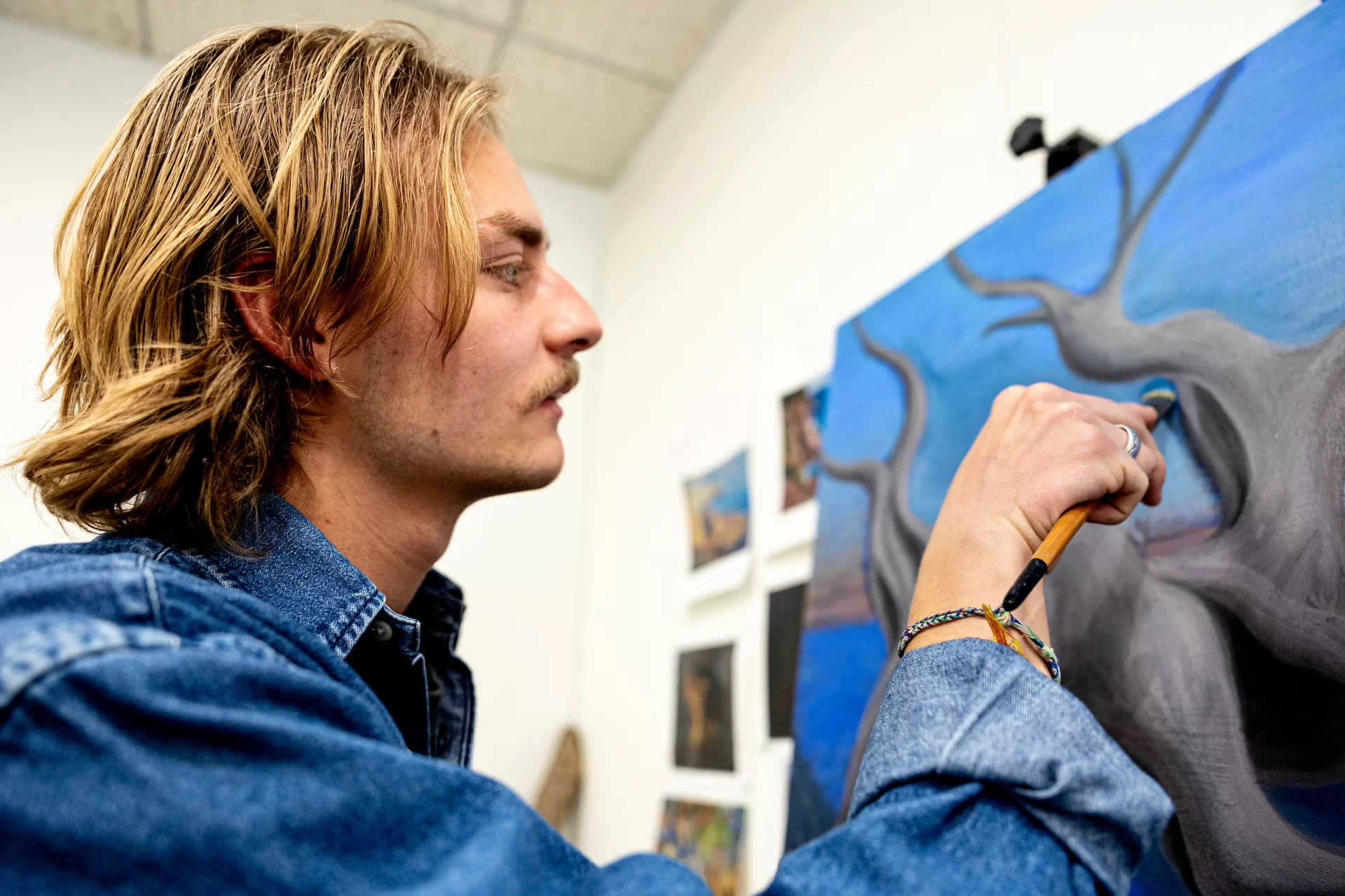 Studio art major George Peck ’24 of Philadelphia, in his Olin Arts Center studio with his paintings for his senior exhibition.

Artist Statement
“This project is an exploration of the way mental images render and fade within my memory. My paintings are of spaces, moments, ideas, and objects that exist within my recollection of a camping trip on the Cutler Coast in northern Maine during the fall of 2023. These paintings are displayed with an array of driftwood that I collected in Maine that same fall. Themes of weathered wood and campfires are both metaphors for memory in this series because of their presence in the experiences I am referencing, and the connections I draw between what I see in their nature and the nature of memories.

The burning campfires in my paintings are a symbol for a moment as it plays out in real time. Campfires are human made, bright, and temporary, just as a lived experience is. The way the fire illuminates parts of the landscape, while other aspects remain in darkness, is similar to how my memories have a clear center from which my perceptions fade.

Driftwood and dead weathered trees can be metaphors for the way something alive, like a moment in space and time, ‘dies’ when the moment has passed. The form of this experience, like a tree, does not disappear, but rather lives on as its own subject, prone to the wind, rain and sun. The wood, like memory, warps, smooths, and sometimes takes on a life of its own, as it is exposed to time and the constant barrage of the elements.

I began collecting natural objects to create sculptures for my thesis in the fall of 2023 and was quickly drawn to driftwood as a material. The beautiful intricacies that come from the natural growth of a tree, followed by the slow wear of the ocean, leads to incredible ‘sculptures’ crafted by the cycle of life and time. I realized that I was drawn to driftwood because of this quality and did not want to alter the pieces I had collected in any significant w