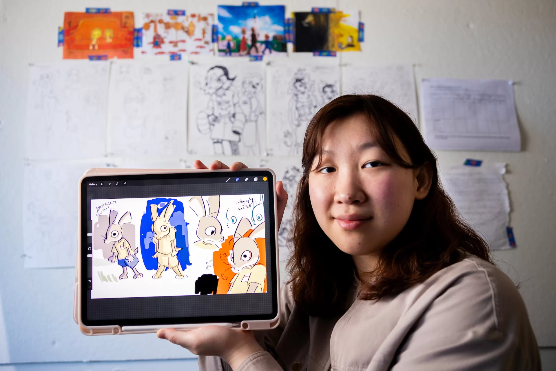 Studio art major Yuri Kim ’24 of East Brunswick, N.J., in her Olin Arts Center studio and a nearby classroom with a white board with her iPad and drawings for her digital animation project on March 8, 2024.

Artist Statement
“In the middle of the woods in western Pennsylvania, a young girl experiences a spiritual encounter that will change her life. In other words, it’s another Good Friday. 

Small communities are a funny thing for a child growing up in one. Sometimes, you are told to do things that you do not particularly understand. Sometimes, you are told that there are certain things that belong to the inside, and other things that belong to the outside. Sometimes, you are taken behind closed doors, and responsible for keeping silent about the things that happen behind them. Sometimes, you’re told that your imagination gets the best of you. Sometimes you agree.

Children interpret such events in fascinating ways. These interpretations are often rebutted, degraded, and dismissed by those around them. Sometimes, this is because the way children interpret things is not seen as particularly appropriate for the occasion. Silliness, weirdness, discomfort, inconsistencies and all – this work embraces these maligned apostles with its arms wide open. It sees the valuable things that lay inside children’s daydreams – eggs, waiting to be hatched. 

This work was made possible through digital animation and compositing. It started from a daydream, then turned into a story. The work parallels my research into the colonial origins of Easter – both in its roots in Europe as well as its start in Pennsylvania. I found repeated violences in the colonization of pagan traditions, the colonization of children’s innocence, and the colonization of the land. I hope you consider these parallels in the viewing of this work.

I hope that you are comforted. I hope that you are discomforted. I hope that you are reminded. I hope that you forget. I hope that you see. I hope y