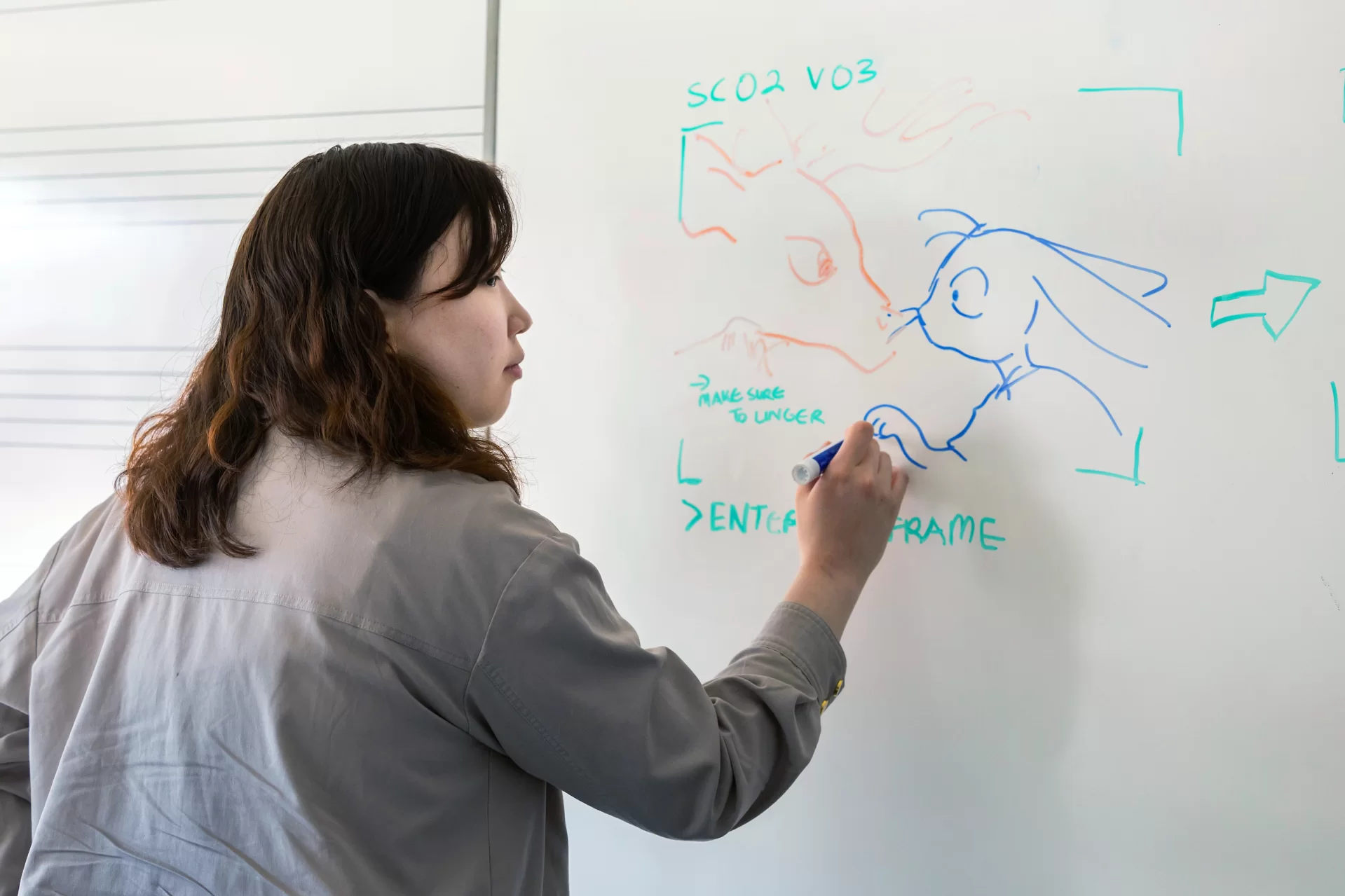 Studio art major Yuri Kim ’24 of East Brunswick, N.J., in her Olin Arts Center studio and a nearby classroom with a white board with her iPad and drawings for her digital animation project on March 8, 2024.

Artist Statement
“In the middle of the woods in western Pennsylvania, a young girl experiences a spiritual encounter that will change her life. In other words, it’s another Good Friday. 

Small communities are a funny thing for a child growing up in one. Sometimes, you are told to do things that you do not particularly understand. Sometimes, you are told that there are certain things that belong to the inside, and other things that belong to the outside. Sometimes, you are taken behind closed doors, and responsible for keeping silent about the things that happen behind them. Sometimes, you’re told that your imagination gets the best of you. Sometimes you agree.

Children interpret such events in fascinating ways. These interpretations are often rebutted, degraded, and dismissed by those around them. Sometimes, this is because the way children interpret things is not seen as particularly appropriate for the occasion. Silliness, weirdness, discomfort, inconsistencies and all – this work embraces these maligned apostles with its arms wide open. It sees the valuable things that lay inside children’s daydreams – eggs, waiting to be hatched. 

This work was made possible through digital animation and compositing. It started from a daydream, then turned into a story. The work parallels my research into the colonial origins of Easter – both in its roots in Europe as well as its start in Pennsylvania. I found repeated violences in the colonization of pagan traditions, the colonization of children’s innocence, and the colonization of the land. I hope you consider these parallels in the viewing of this work.

I hope that you are comforted. I hope that you are discomforted. I hope that you are reminded. I hope that you forget. I hope that you see. I hope y