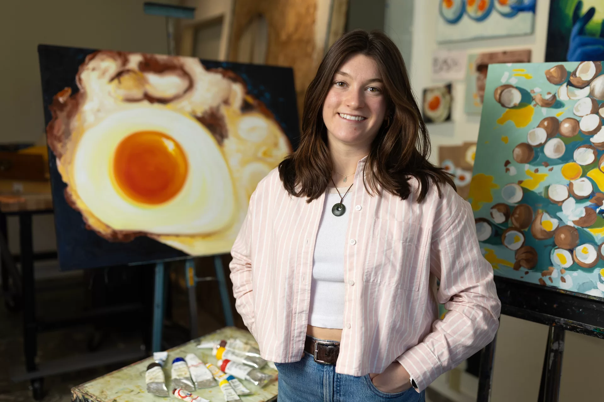Studio art major Avery Mathias ’24 of Needham Heights, Mass., in her Olin Arts Center studio with her paintings for her senior exhibition.

Artist Statement
“My body of work seeks to focus on the mundane as a worthy subject matter to explore light, color, and the beauty in the ordinary. I have focused on a single subject—the chicken egg— as it is a universally recognizable object that is often overlooked. Given that the chicken egg is so common, it is accompanied by a variety of connotations that the audience can examine with the work. Combined with the striking contrast of the yellow-orange yolk with egg whites, the chicken egg encapsulates the concept of finding intrigue and beauty in the mundane.

In order to emulate traditional still life painting, I stretched and gessoed the canvases by hand and used oil paint as my medium. While I was inspired by historical still lives by female artists such as Gluck (Hannah Gluckstein), Ethel Sands, and Vanessa Bell, I was also influenced by the modern still life painter Leah Gardner. She is a young, self-taught artist whose work consists of a series of common objects captured with bright colors on a plain background. Her use of light and color inspired me to focus on daily life and the functioning of seemingly insignificant mechanisms which led to my involvement with biology and cooking.

I have particularly fond memories of making breakfast with my father on the weekends as a kid and enjoy food and how a shared meal brings people together. While food and people’s relationship with it comes with a range of emotions and connotations, everyone can recognize and connect to the symbol of a fried egg. In addition to providing valuable nutrition, an egg can symbolize or invite other associations such as life and sexuality. The lack of context included in my work invites the audience to bring their own associations and significance to each piece. The egg is also the epitome of routine as a chicken lays one egg every day a