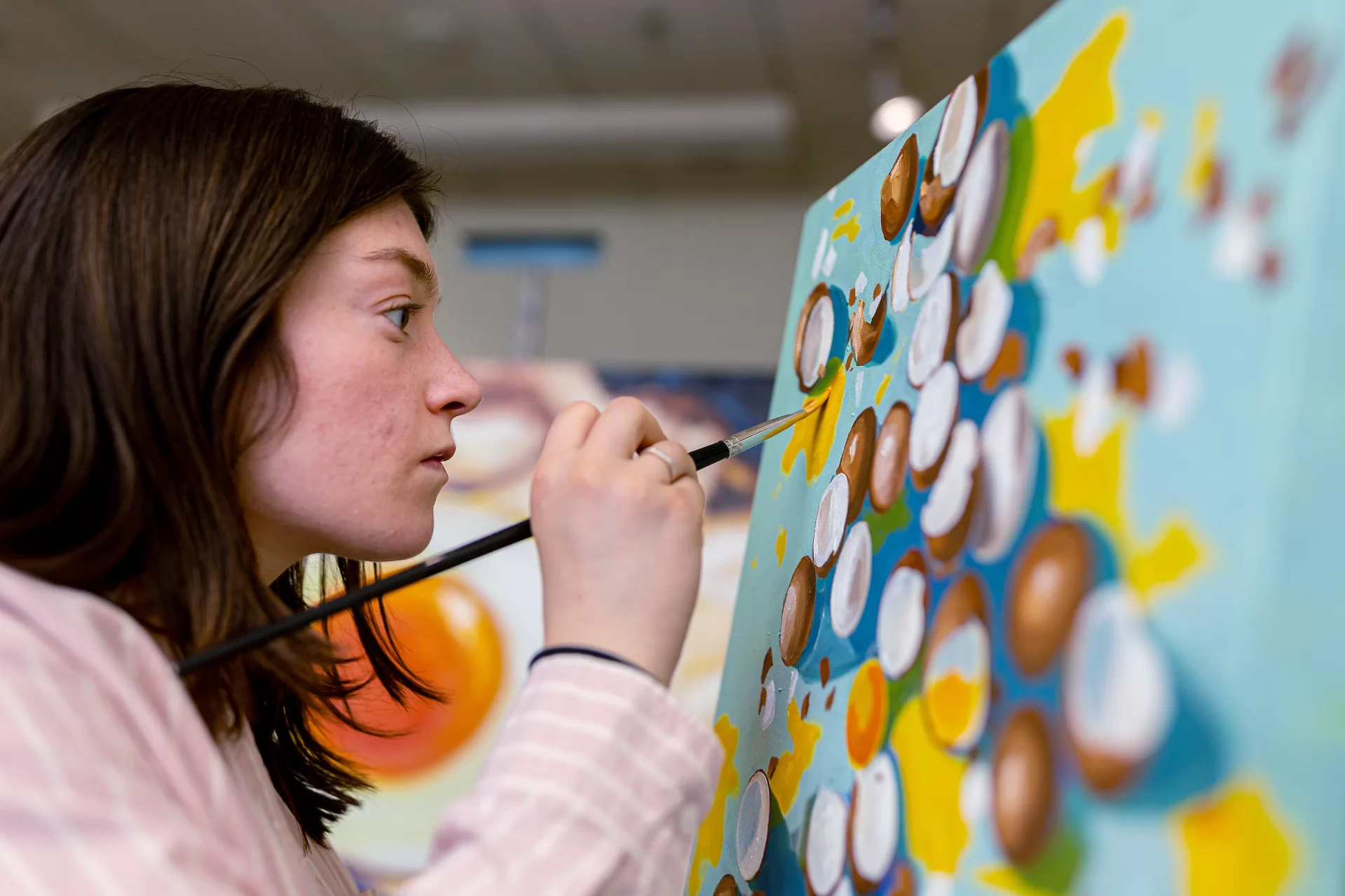 Studio art major Avery Mathias ’24 of Needham Heights, Mass., in her Olin Arts Center studio with her paintings for her senior exhibition. Artist Statement “My body of work seeks to focus on the mundane as a worthy subject matter to explore light, color, and the beauty in the ordinary. I have focused on a single subject—the chicken egg— as it is a universally recognizable object that is often overlooked. Given that the chicken egg is so common, it is accompanied by a variety of connotations that the audience can examine with the work. Combined with the striking contrast of the yellow-orange yolk with egg whites, the chicken egg encapsulates the concept of finding intrigue and beauty in the mundane. In order to emulate traditional still life painting, I stretched and gessoed the canvases by hand and used oil paint as my medium. While I was inspired by historical still lives by female artists such as Gluck (Hannah Gluckstein), Ethel Sands, and Vanessa Bell, I was also influenced by the modern still life painter Leah Gardner. She is a young, self-taught artist whose work consists of a series of common objects captured with bright colors on a plain background. Her use of light and color inspired me to focus on daily life and the functioning of seemingly insignificant mechanisms which led to my involvement with biology and cooking. I have particularly fond memories of making breakfast with my father on the weekends as a kid and enjoy food and how a shared meal brings people together. While food and people’s relationship with it comes with a range of emotions and connotations, everyone can recognize and connect to the symbol of a fried egg. In addition to providing valuable nutrition, an egg can symbolize or invite other associations such as life and sexuality. The lack of context included in my work invites the audience to bring their own associations and significance to each piece. The egg is also the epitome of routine as a chicken lays one egg every day a