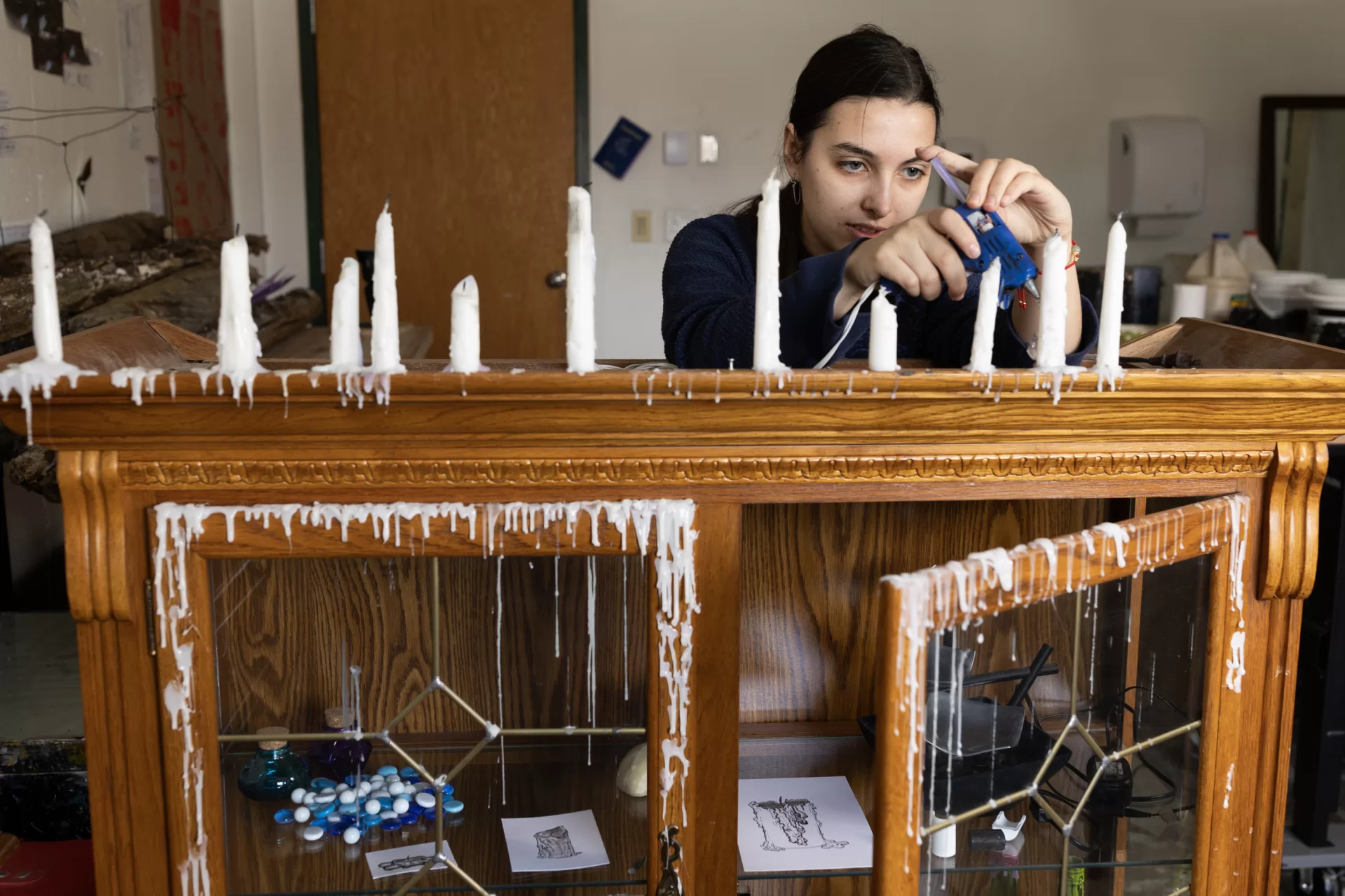 Olivia Rabin ’24 of Montclair, N.J., works in her Olin Arts Center studio on March 21, 2024. From a young age, I was fascinated by nature and the fantastical. This and the works by people similarly inspired by the natural world inspire my current work. While I am interested in many different things, I am always working to visualize them to help me understand how I connect them internally. In my work, I am trying to synthesize my own process into something tangible and observable. I am exploring the connections between my headspace, the act of expression, and the physical world. I want to explore the emotions and sensations of the world around me, being captivated by nature and the fantastical. I love the mysterious blues and otherworldly qualities of water, especially found in oceans and waterfalls. I am thoroughly enchanted by them. This was only heightened by my favorite creative works like The Blue Planet by David Attenborough. This film not only allowed me to see the ocean’s depths in ways I had never seen before but also showed me ways in which form can create fantastical emotions out of the real. Works like Hayao Miyazaki’s Laputa: Castle in the Sky expand on this in artistic and sensory ways that become their own mythology. Works by artists like Heikala and Gabriel Picolo expand upon mundane elements of reality, abstracting them into the fantastical. Heikala’s illustrations often explore the magic in mundane moments by abstracting an element like size, time, or location to convey an emotion fantastically. In Picolo’s Icarus and the Sun, the already metaphorical wax of Icarus’ wings becomes more emotional as Icarus’s body is made of wax and the sun becomes his lover. Wax is quite captivating for me as its qualities are intrinsically related to water. Wax flows like water but as it cools and solidifies it almost freezes a moment in time and space allowing for the magic to be captured. In these works, I was able to find connections between my interests in nature, magic, water, and wax. The emotions I feel from these are something that I both want to explore and give back. I often struggle with the translation of ideas to form. I want each work to be expressed in the medium best suited for it. As my work is driven by emotions and physical sensations, I frequently work based on intuition. That being said, this process is not without planning as I often work on mock versions on a smaller scale. Sometimes these versions then end up becoming part of the work, if not the work itself. The liquid qualities of watercolor, gouache, and ink are sensorily interesting and a logical way for me to convey fluidity which is important to me. The nature of these materials afford me the ability to work in layers which I like to employ in my work. I am interested in illustrative and abstract work that is rooted in reality while distorting it or finding new meanings. The same ideas in the conversation around film, with its ability to capture some version of reality while also being an abstraction, also resonate with me. I see parallels in physical and digital mediums’ ability to collect objects or aspects of reality while constructing spaces beyond our physical world. The idea of collection and storage as a method of constructing, warping, and destroying meaning is key to my artistic practice. It reminds me of my interests in shelves and storage spaces and how they can create new meanings or sites of connection. These art practices become a physical manifestation of these ideas. I consider my work experimental, as I try new things that are based on an internal dialogue and database. My art becomes an expression of this dialogue as I attempt to make sense of things that barely make sense to me. I explore how these things are stored in my head like objects of wonder on a shelf.