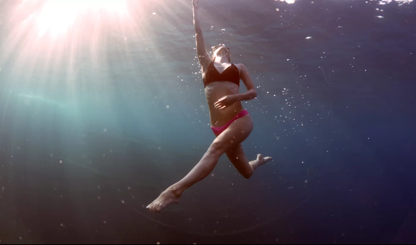 A woman dances underwater as sunlight filters through the water from above.