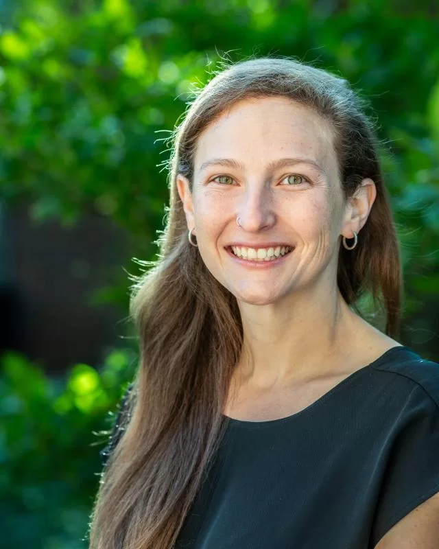 A woman smiles in front of greenery.