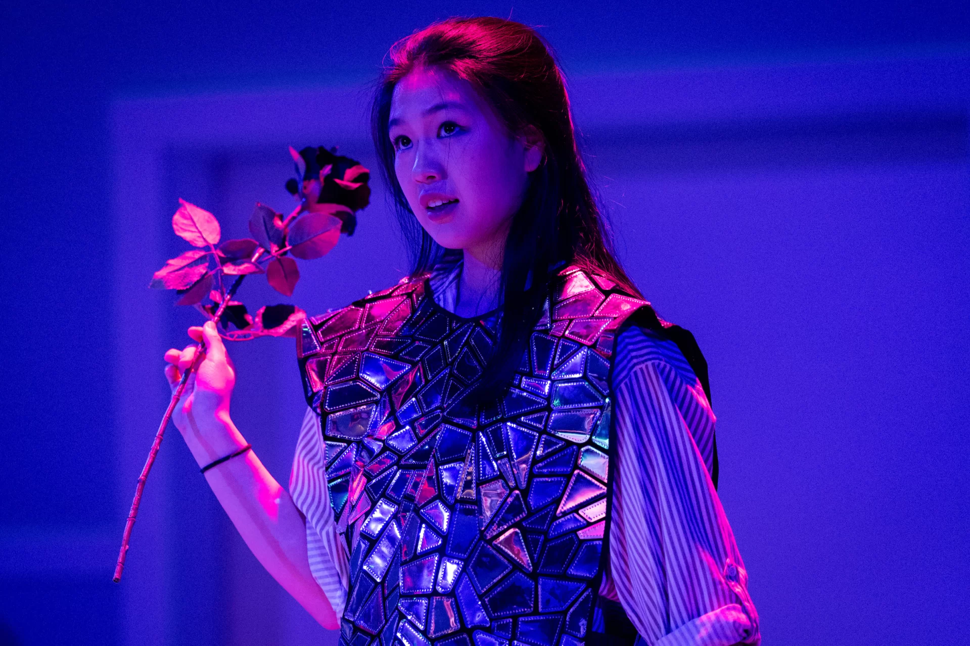 A woman holds a flower onstage beneath pink lights.